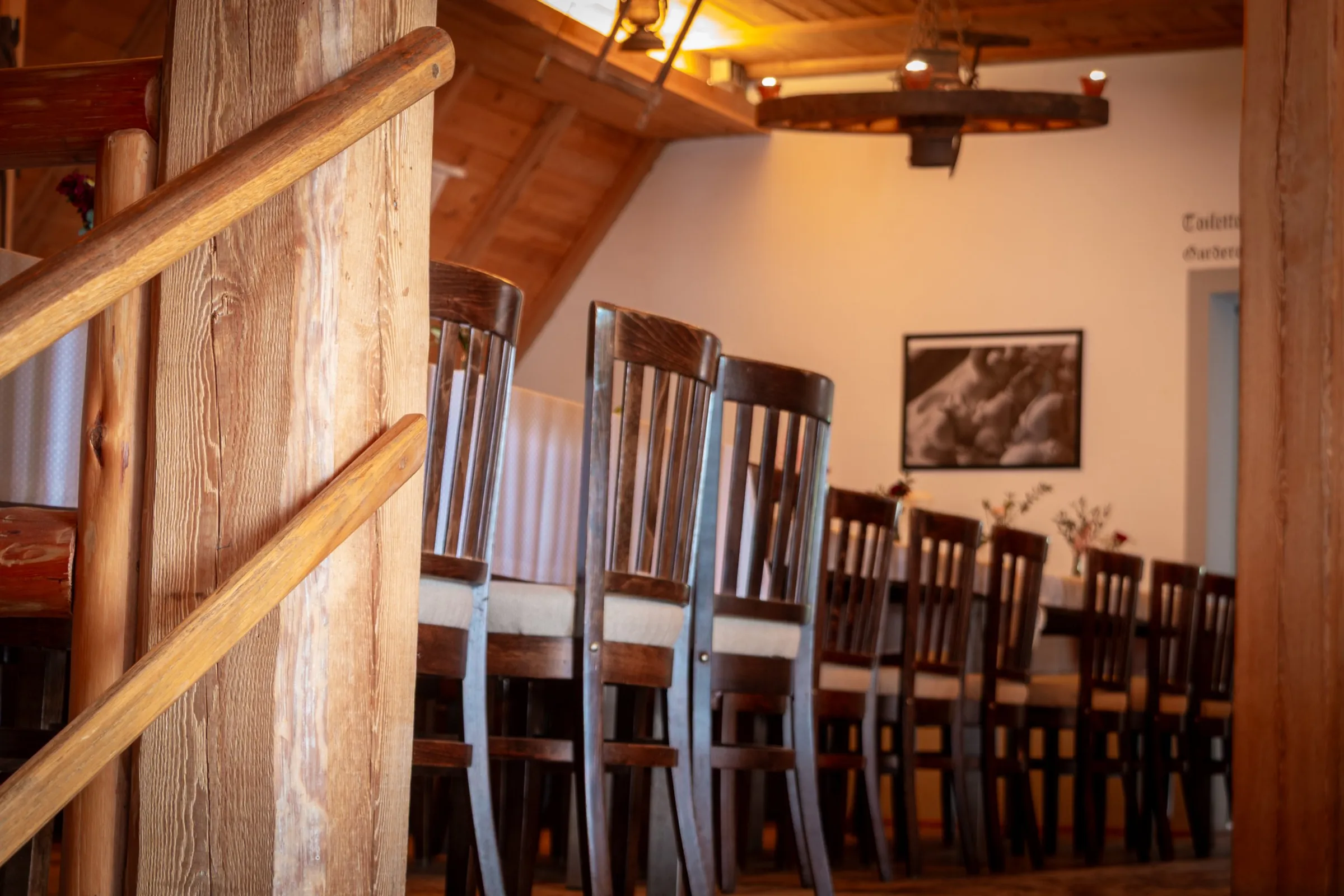 Stuhlreihe an der langen Tafel im Stodl, vorne der Holzpfosten der Treppe, darüber ein Wagenrad als Leuchter
